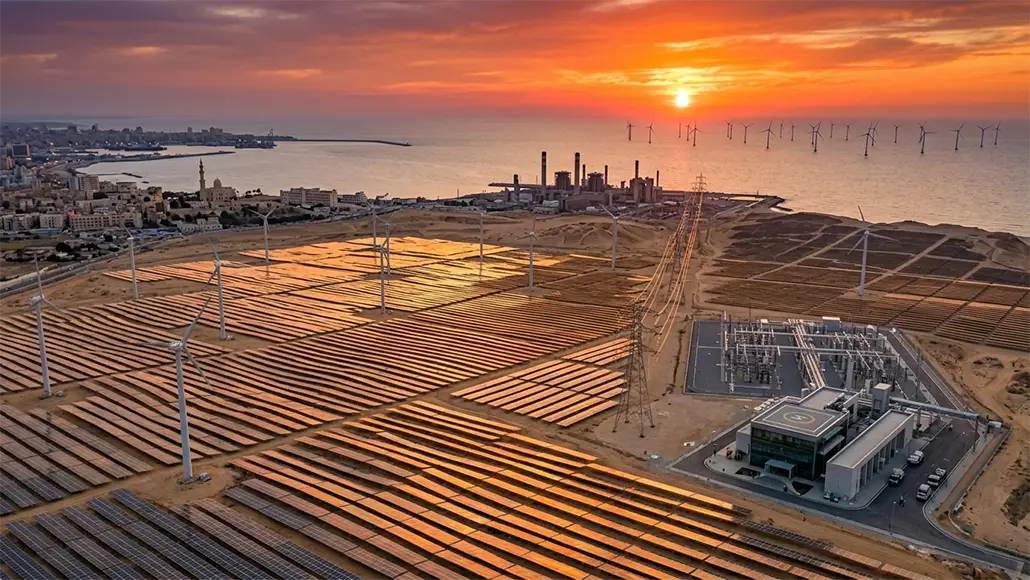 Sunset over a coastal solar farm with wind turbines and offshore wind farms in the distance, beside an industrial plant.