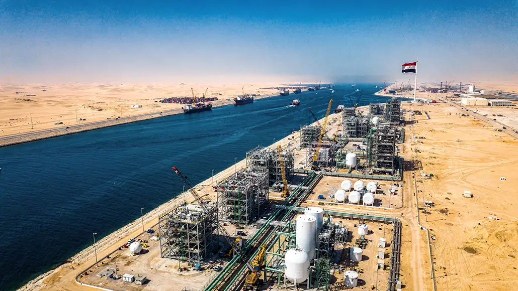 Aerial view of an oil and gas processing facility along a desert canal, with towers, tanks, cranes, and an Egyptian flag.