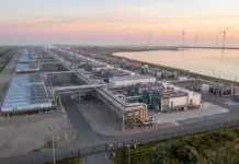 Poland, Japan, Hungary Ink 2 Hydrogen, Green Ammonia Deals Industrial hydrogen production facility with extensive piping and cylindrical tanks labeled H2 by a coastal shoreline, wind turbines visible in the distance.