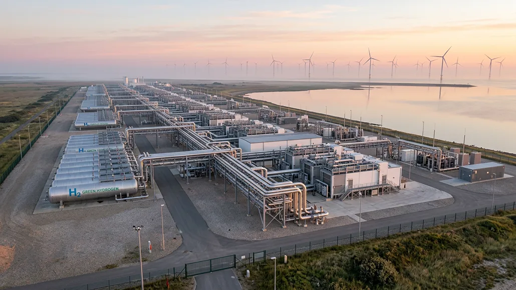 Industrial hydrogen production facility with extensive piping and cylindrical tanks labeled H2 by a coastal shoreline, wind turbines visible in the distance.