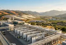 €6bn Approval for Renewable Hydrogen Production in Italy Renewable energy facility with extensive pipelines, storage tanks, and a modern building set in rolling hills with wind turbines in the distance.