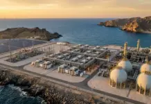 800-1,000MW Green Hydrogen for Power Generation Oman Tender Industrial complex with large spherical storage tanks and piping on a rocky coastline at sunset by the sea.