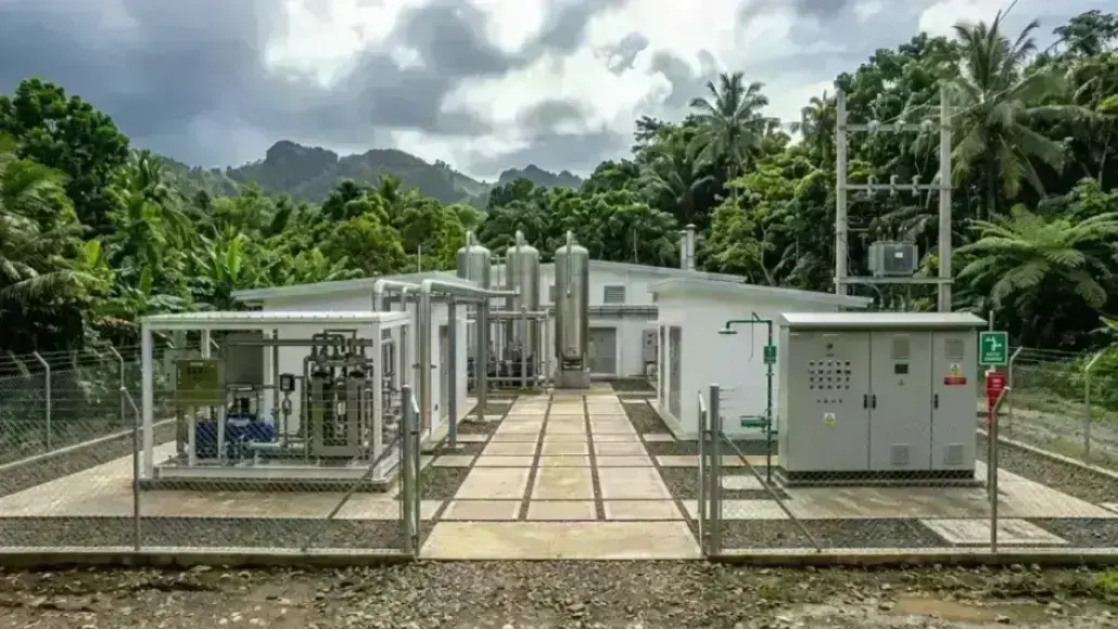 Outdoor industrial facility with silver tanks and machinery inside a chain-link fence, set in a tropical, tree-filled area.