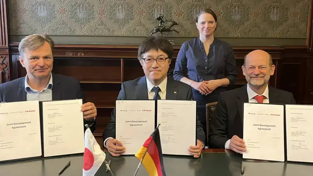 Three executives sign a Joint Development Agreement at a formal signing ceremony, with Japan and Germany flags on the table and a woman standing behind them.