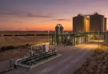 Push For Hydrogen and Biomethane Strategies in Spain Industrial gas plant at sunset with tall silos, complex piping, and wind turbines on the horizon.