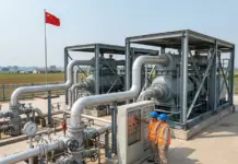 First 1mn-Cubic-Mtr Salt Cavern Hydrogen Storage in China Two technicians in hard hats and safety vests operate a control panel beside a network of large insulated pipes at an industrial facility, with a Chinese flag in the background.
