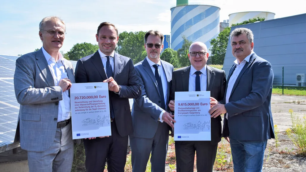 Five men in suits stand outdoors, holding two large certificates showing 20,720,000 euros and 9,615,000 euros, with solar panels and storage tanks in the background.