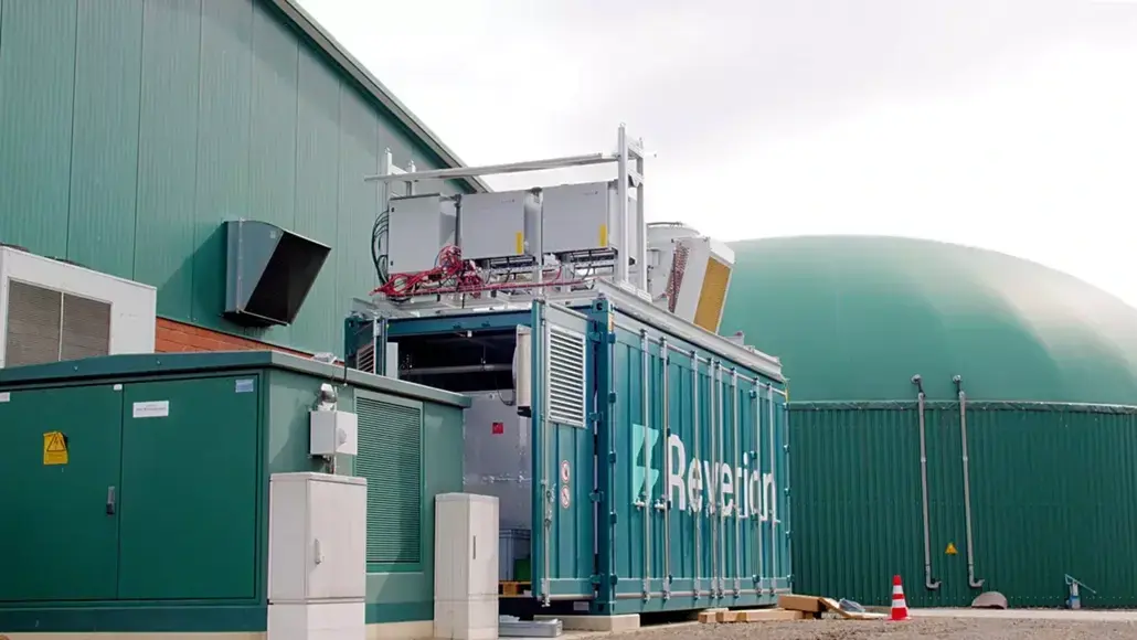Industrial equipment setup beside a green building; a blue container with branding and exposed electronics on top.