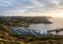 Advancement In Avalon Isthmus, The Wind-To-Hydrogen Project Coastal industrial site with wind turbines on rolling hills overlooking the sea and a docked cargo ship nearby.