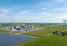 Advancing Large-Scale Denmark Renewable Hydrogen Project Renewable energy site with solar panels in the foreground, a processing plant, wind turbines on the horizon, and a hydrogen (H2) sign to the right.