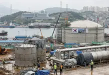 Korea Eyes Domestic Liquefied Hydrogen Storage Infra Harbor-side construction site with large cylindrical tanks, pipes, cranes, and workers in safety gear near the water.