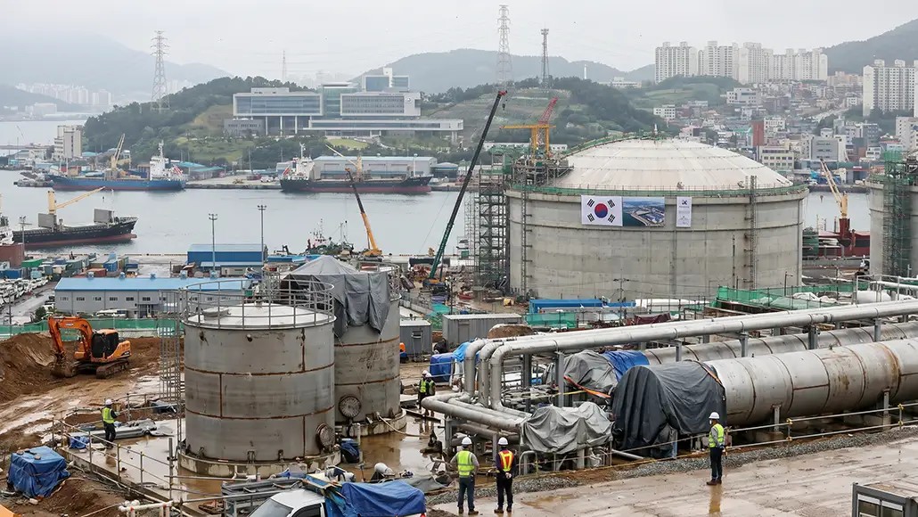 Harbor-side construction site with large cylindrical tanks, pipes, cranes, and workers in safety gear near the water.