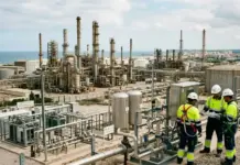 Efforts to Push Green Hydrogen Production in Spain Refinery Industrial refinery complex with tall processing towers and extensive piping; workers in hard hats and high-visibility gear reviewing plans at the site.