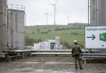 Eneos May Import Green Hydrogen-Based e-Fuels from Germany Industrial hydrogen production site with large storage tanks, piping, and a worker in protective gear; wind turbines on green hills in the background.