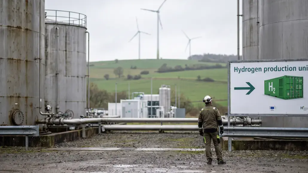 Industrial hydrogen production site with large storage tanks, piping, and a worker in protective gear; wind turbines on green hills in the background.