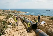 Hydrogen-Ready Pipeline Project in Malta Supported by EU Construction workers in safety vests and helmets install a large pipeline along a rocky coastal site by the sea.