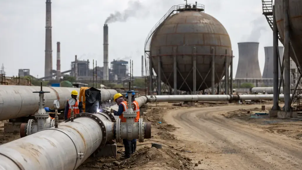 Workers in orange vests and yellow helmets weld and inspect a large outdoor oil pipeline.