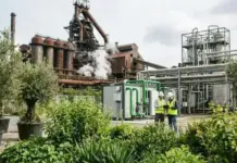 Green Molecules to Green Steel Platform Introduced Two workers in hard hats and high-visibility vests inspect equipment at an industrial plant with green piping and smokestacks outside. (informative)